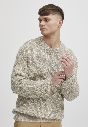 Beiger Strickpullover mit strukturiertem Muster, gerippten Bündchen und runder Ausschnitt, getragen von einem Modell mit kurzem Haar vor neutralem Hintergrund.