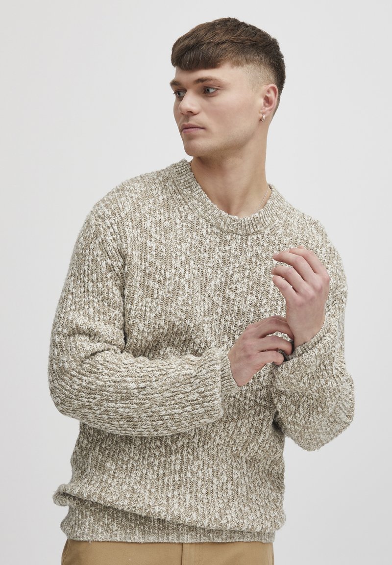 Beige knit sweater with a textured pattern, ribbed cuffs, and crew neckline, worn by a model with short hair in a neutral background.