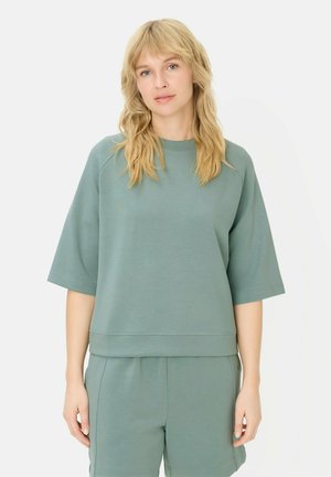 Kobieta o blond włosach, ubrana w luźną, szałwiowo zieloną bluzkę z krótkim rękawem i pasujące do niej szorty, stojąca na tle białego tła.