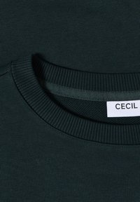 Mörkgrön sweatshirt med ribbad halsringning och en slät textur. Etiketten säger "CECIL" och är placerad nära halsens söm.