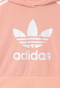 Haut à capuche rose clair avec un logo trefoil blanc Adidas et le nom de la marque sur la poitrine, en tissu texturé et avec des détails de bord contrastés.