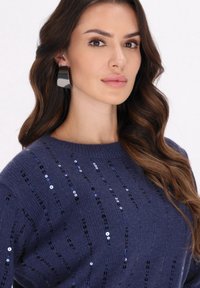 Pulover marin cu dungi verticale din paiete; textură moale, decolteu tip crew. Se poartă cercei mari, lucioși, de culoare gri, adăugând un detaliu îndrăzneț.