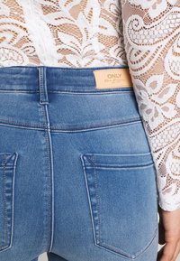 Gros plan sur une personne portant un jean en denim bleu taille haute avec de grandes poches arrière et un haut en dentelle blanche à motifs floraux.