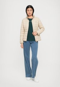 Beige pufferjack met een staande kraag, gecombineerd met een donkergroen shirt en blauwe flared jeans. Gladde textuur, eenvoudig ontwerp, gouden rits.