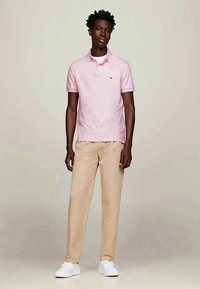 Polo shirt rosa chiaro con colletto, abbinato a pantaloni chinos beige e sneakers bianche. Design semplice con un logo in evidenza sulla maglietta.