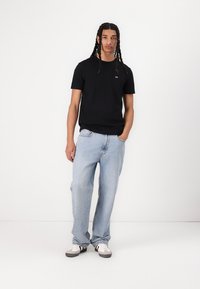 T-shirt en coton noir à col rond, associé à un jean ample bleu clair. Le modèle se tient debout avec une main dans la poche, portant des baskets blanches.