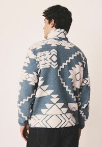 Fleece pullover in blue with white geometric patterns, featuring a high collar and textured, plush fabric. Visible back details and seams.