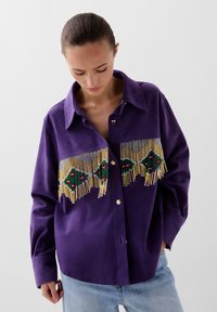 Camicia di jeans viola con un design frangiato e dettagli di perline multicolori. Tessuto morbido e liscio con colletto classico e vestibilità ampia.
