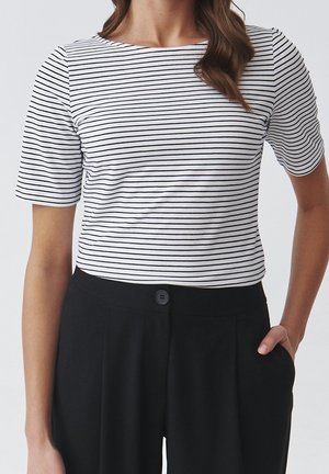 Mujer con una camiseta blanca de manga corta con finas rayas horizontales negras y pantalones negros de talle alto, una mano en el bolsillo.