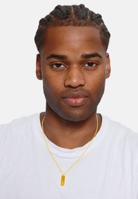 Goldkette mit einem rechteckigen Anhänger an einer dünnen Kette, getragen mit einem weißen T-Shirt. Der Hintergrund ist schlicht weiß.