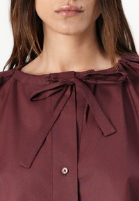 Maroon button-up shirt with a rounded neckline, tie detail, and light texture. Features a central button and gathered fabric at shoulders.