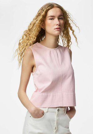 Ung kvinne med langt krøllete blondt hår iført en ermeløs lyserosa topp og hvite bukser, med hendene i lommene, stående mot en hvit bakgrunn.