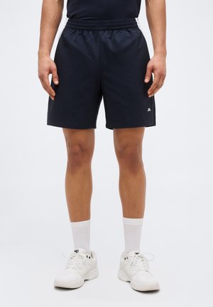 Mann trägt schwarze Shorts mit elastischem Bund, weiße Sportsocken und weiße Sportschuhe, steht vor hellem Hintergrund.