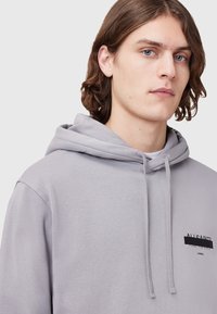 Lichtgrijze hoodie met een voorzak en een trekkoord. Heeft een klein zwart logo. Zachte katoenen stof met een ontspannen pasvorm.