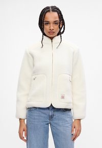 Giacca in pile crema con colletto alto, due tasche frontali e zip frontale. Presenta una patch con logo all'orlo. Indossata sopra jeans blu.
