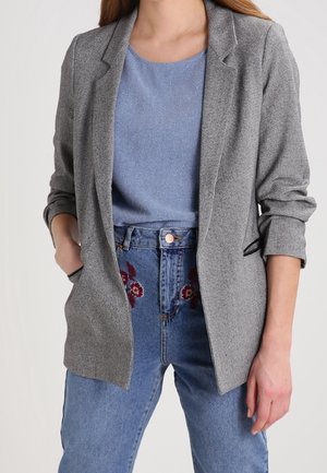 Femme portant un blazer gris sur un haut bleu et un jean bleu taille haute avec une broderie florale rouge, mains dans les poches du blazer.