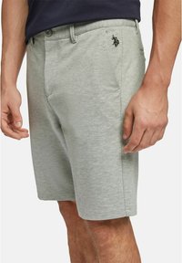 Homme portant un short gris clair longueur genou avec un petit logo noir sur la poche droite et une chemise foncée rentrée à l’intérieur.