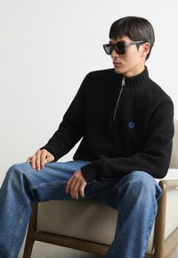 Maglione nero a maglia con colletto alto e zip argentata, abbinato a jeans azzurri, seduto su una sedia di legno. Dettaglio semplice del logo.