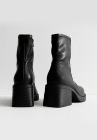 Par de botas de cuero negro hasta el tobillo con tacones gruesos, costuras visibles y cremallera trasera, mostradas desde la parte trasera sobre una superficie blanca.
