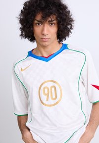 Camiseta de fútbol blanca con acentos en verde, azul y rojo. Presenta un patrón de cuadrícula texturizada, un emblema dorado con el número "90" y un logo de Nike.