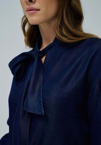 Blusa azul marino con un detalle de lazo en el collar, que presenta una textura suave y un corte relajado. La tela parece ligera y ligeramente brillante.