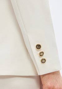 Manche de blazer couleur crème avec trois boutons décoratifs noirs et dorés près du poignet sur le bras d'une personne.