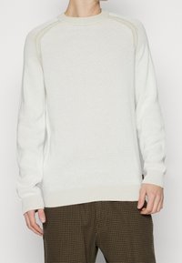 Jasnobeżowy sweter z dzianiny z prążkowanymi mankietami i krawędzią, ozdobiony teksturowanym wzorem. W zestawie z brązowymi spodniami w kratkę.