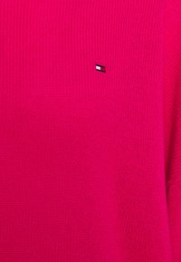 Pull rose en maille avec une texture côtelée et un logo brodé en noir, blanc et rouge sur le côté gauche.