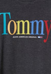 Camiseta gris con "Tommy" en letras multicolores (azul, amarillo, naranja) encima de "JEANS AMERICAN ORIGINAL" en blanco, con un acento de rayas de colores.