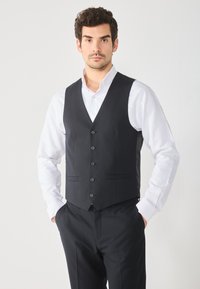 Gilet noir en tissu lisse, avec un d�écolleté en V, six boutons et deux poches avant, porté sur une chemise blanche. Coupe ajustée.