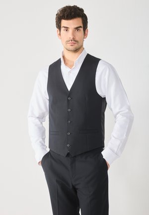 Black vest made of smooth fabric, featuring a V-neck, six buttons, and two front pockets, worn over a white shirt. Tailored fit.