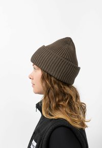 Brown ribbed knit beanie with a peaked top, worn over wavy hair, in profile against a white background. Material appears soft and textured.