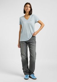 T-shirt azzurro chiaro a V realizzato in tessuto morbido, con maniche corte, che presenta un piccolo logo sul petto, abbinato a jeans grigi e sneakers blu.