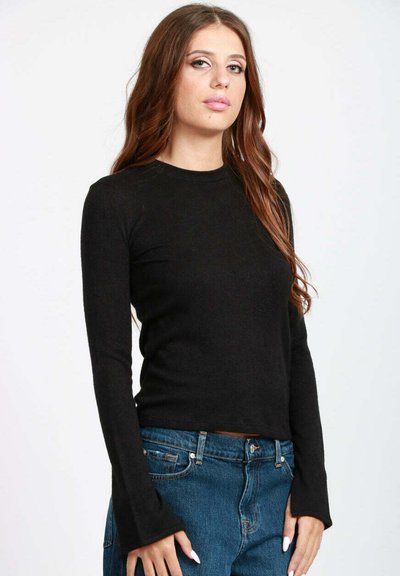 Giovane donna con lunghi capelli castani, indossa un top a maniche lunghe nero e jeans blu, in piedi davanti a uno sfondo bianco semplice.