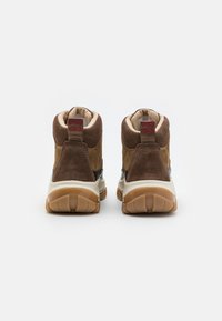 Brązowe zamszowe sneakersy w średnim fasonie z wyściełanym kołnierzem, gumową podeszwą i zaokrąglonym kształtem. Elementy charakterystyczne to kontrastowa ciemniejsza panel i detal z logo.