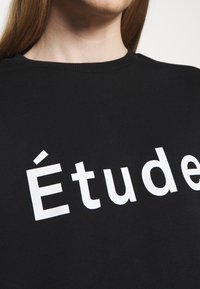 Sudadera negra con cuello redondo que presenta un gran texto blanco que dice "Étude". La tela parece suave, con textura mínima.