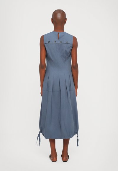 Mėlynas suknelė be rankovių su klostuotu dizainu, rakto skylutės formos nugaros detale ir reguliuojamu raišteliu apdailoje. Pagaminta iš minkštos, lengvos medžiagos.