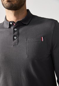 Polo de manga larga gris oscuro hecho de tela texturizada. Presenta una tapeta de tres botones y un pequeño bolsillo en el pecho con rayas de acento rojas y blancas.