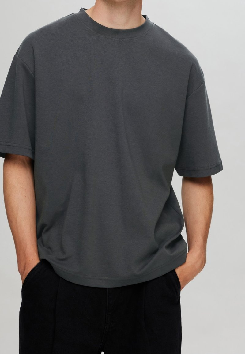 T-shirt en coton gris avec un col rond, des épaules tombantes et des manches courtes. Texture lisse ; coupe décontractée. Porté avec un pantalon noir.