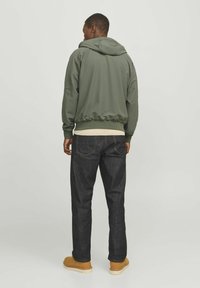 Sudadera con capucha de un verde oscuro, con un dobladillo y puños acanalados, llevada sobre una prenda de color claro, combinada con jeans de mezclilla oscuros y zapatos marrones.