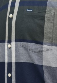 Kockás ing sötétkék, szürke és fehér árnyalatokban, gyöngygombokkal és egy bal oldali mellzsebből, amelyen logó címke található. Pamut anyag.