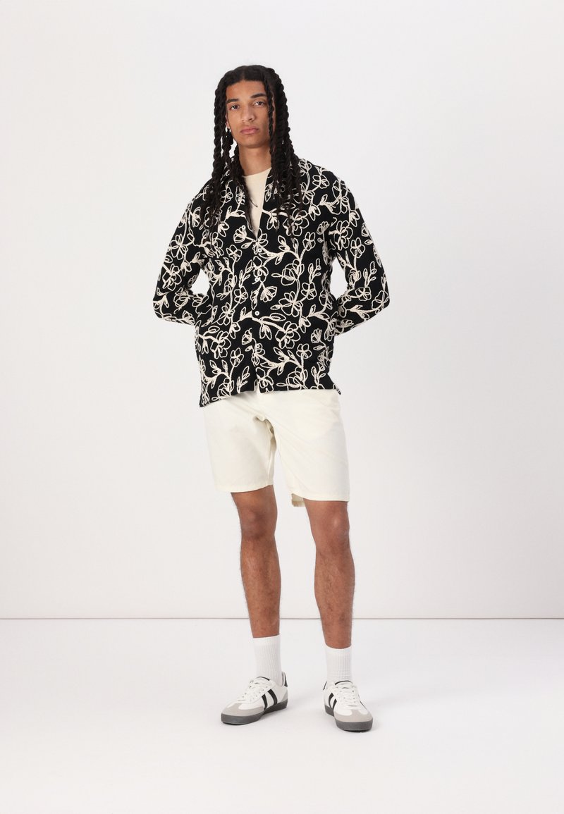 Black jacket with white floral pattern, cream shirt, cream shorts, and black and white sneakers. Model has long braids and a neutral expression.