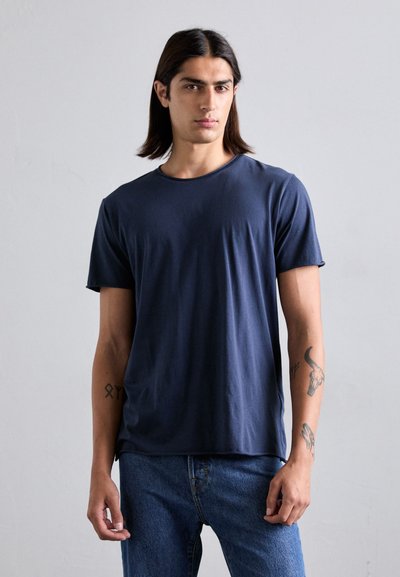 Junger Mann mit langen dunklen Haaren, der ein einfaches navyblaues T-Shirt und blaue Jeans trägt, vor einem schlichten hellen Hintergrund.