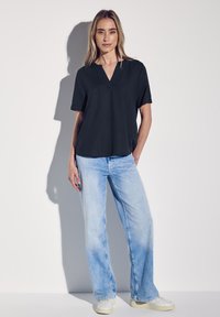 Schwarzes kurzärmliges Oberteil mit V-Ausschnitt, kombiniert mit hellblauen weiten Jeans und weißen Sneakers. Glatte Stoffqualität, lässige Passform.