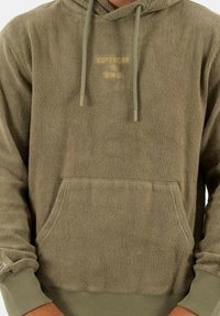 Sudadera con capucha de forro polar verde, con superficie texturizada, capucha ajustable, bolsillo frontal y logo dorado de "Superdry" en el pecho. Los puños son acanalados.