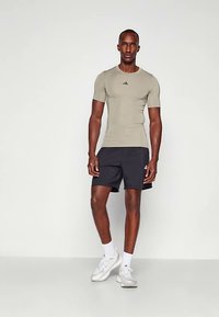 Vaaleanharmaa, tiukan mallinen lyhythihainen urheilupaita mustalla logolla, yhdistettynä mustiin shortseihin. Mallilla on valkoiset urheilukengät ja sukat.