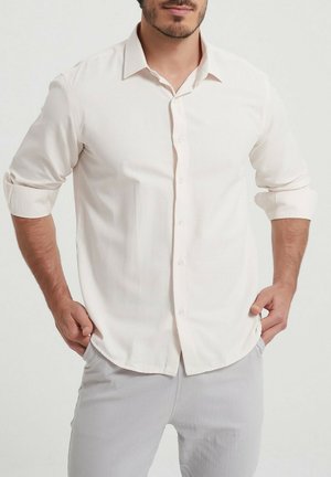 Chemise boutonnée beige clair avec un col pointu, manches longues retroussées aux poignets, fabriquée dans un tissu lisse avec une texture subtile.