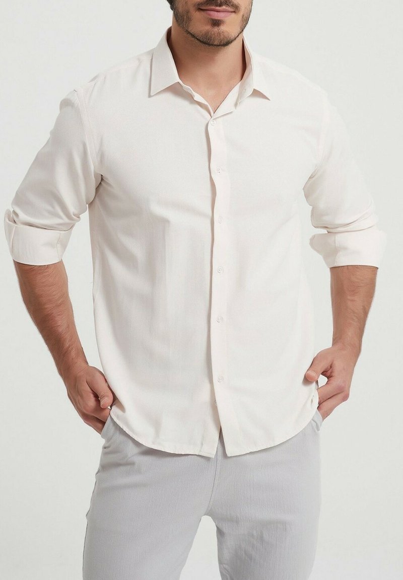 Chemise boutonnée beige clair avec un col pointu, manches longues retroussées aux poignets, fabriquée dans un tissu lisse avec une texture subtile.