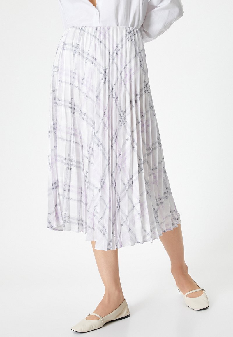 Jupe plissée en tissu blanc translucide avec un motif écossais violet et gris. Taille élastique et coupe longue jusqu'aux chevilles. Associée à des ballerines beiges.