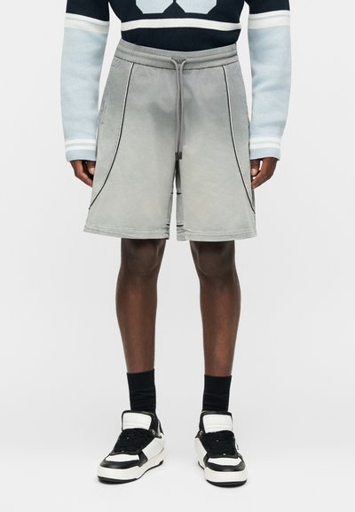 Άνδρας που φοράει ανοιχτό γκρίζο σορτς μέχρι το γόνατο με μαύρο περίγραμμα, μαύρες κάλτσες, μαύρα και λευκά αθλητικά παπούτσια και μπλε σκούρο και ανοιχτό μπλε μακρυμάνικο πουλόβερ.